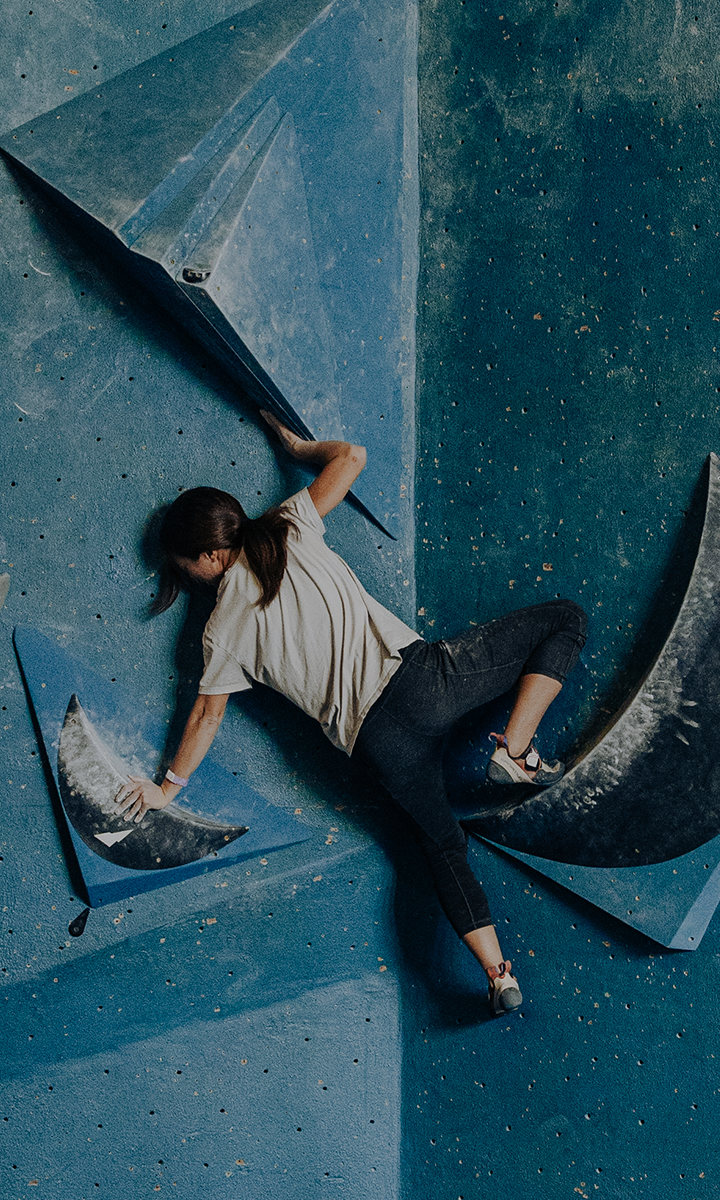 A woman climbs an indoor bouldering wall with large blue geometric holds at Seattle Bouldering Project, showcasing the excitement and Membership Benefits available as she reaches with her left hand while balancing on the wall.