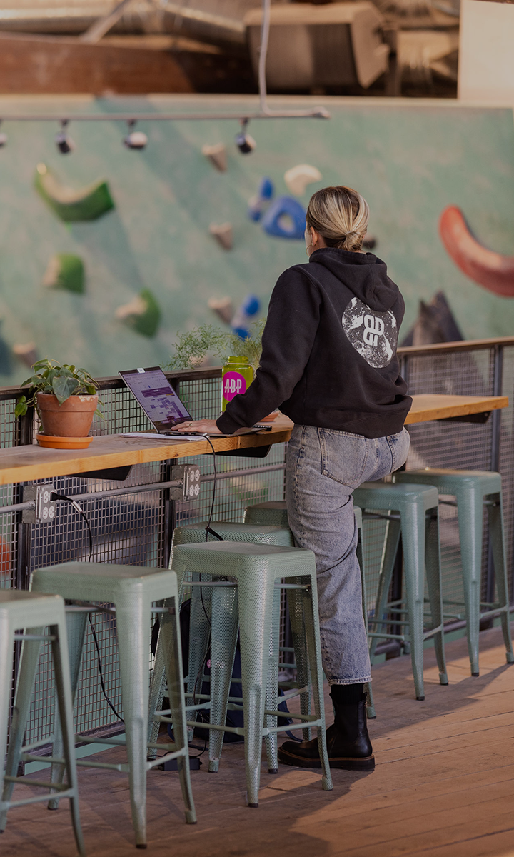 A person with blonde hair sits at a high counter on a green stool, working on a laptop. There are potted plants on the counter, and the Membership Benefits at Seattle Bouldering Project are showcased by the rock climbing wall in the background.