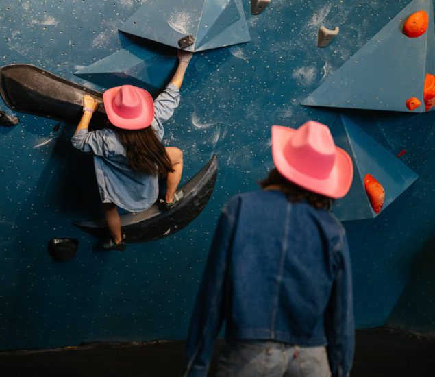 Two people wearing pink cowboy hats and denim jackets; one climbs an indoor rock wall while the other stands nearby, watching. The wall, with blue and orange holds, sets the scene for a lively Seattle climbing competition.