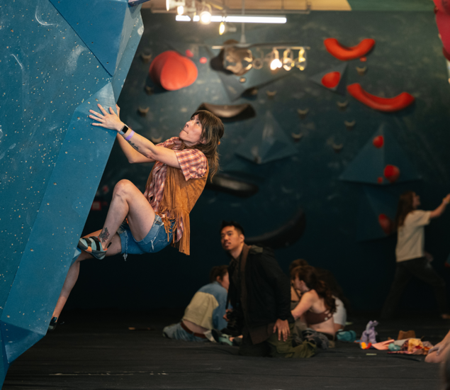 A woman in a fringed vest and denim shorts climbs a blue indoor bouldering wall at a Seattle climbing competition, focused and gripping tightly. Other climbers and spectators are visible in the background.