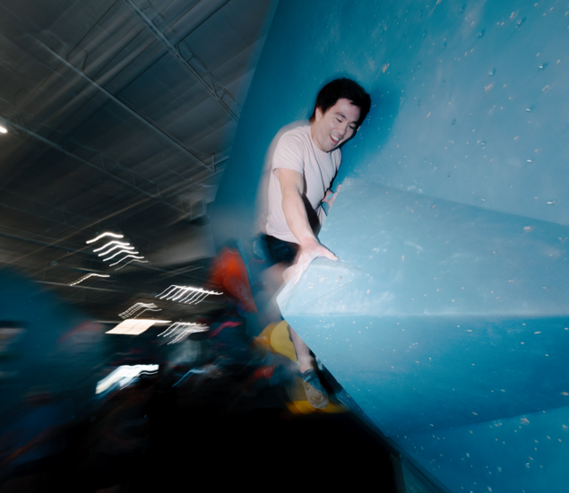 A person in a white shirt climbs an indoor blue bouldering wall, gripping a large hold with one hand; the blurred background emphasizes motion and intensity—perfectly capturing the spirit of Climb for Community.