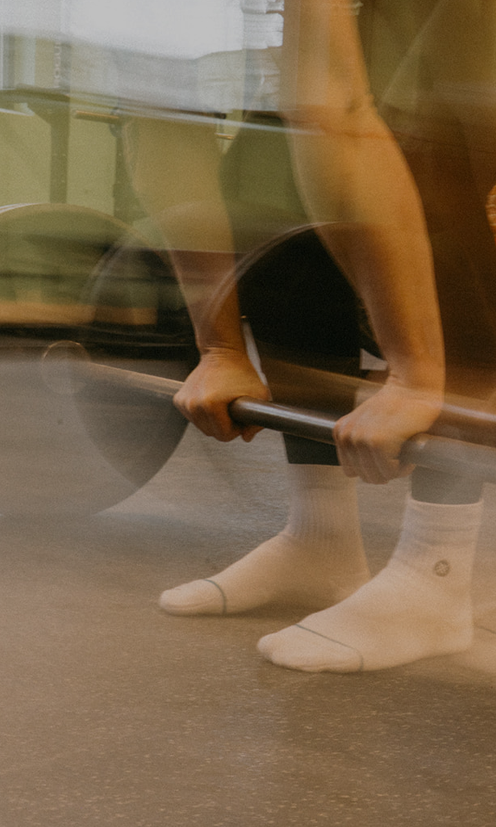 A person wearing white socks prepares to lift a barbell in a gym, shown from the knees down, with a blurred effect capturing motion—much like the dynamic Membership Benefits at Seattle Bouldering Project keep you moving forward.