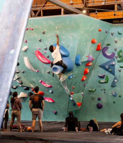 A climber scales an indoor bouldering wall covered in colorful holds as several people watch from padded flooring, enjoying the Membership Benefits at Seattle Bouldering Project in this bustling climbing gym.
