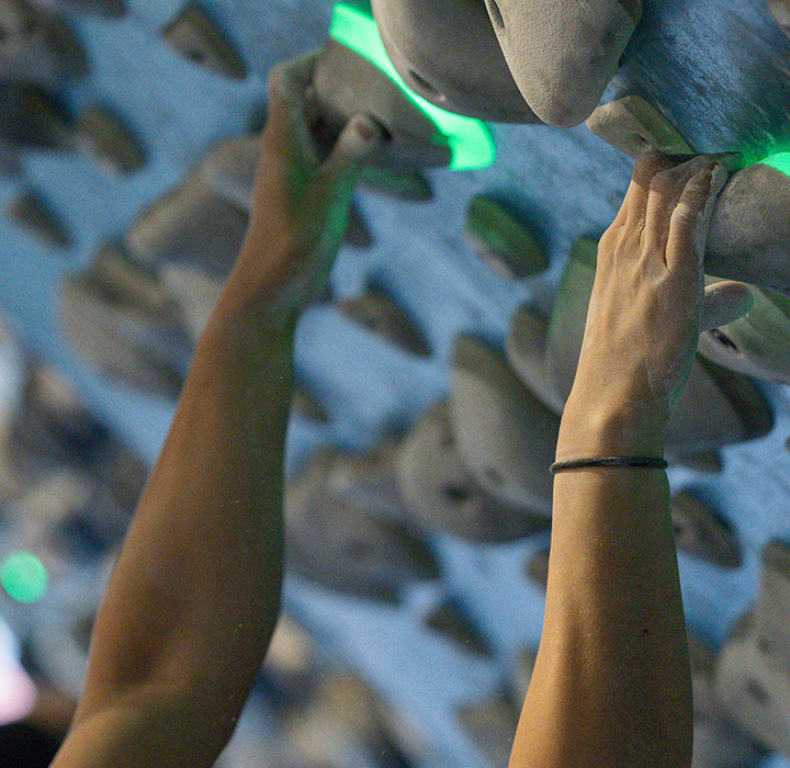 Close-up of people climbing an indoor rock wall, focusing on hands gripping illuminated green climbing holds and poplar training boards, with blurred climbers and holds in the background.