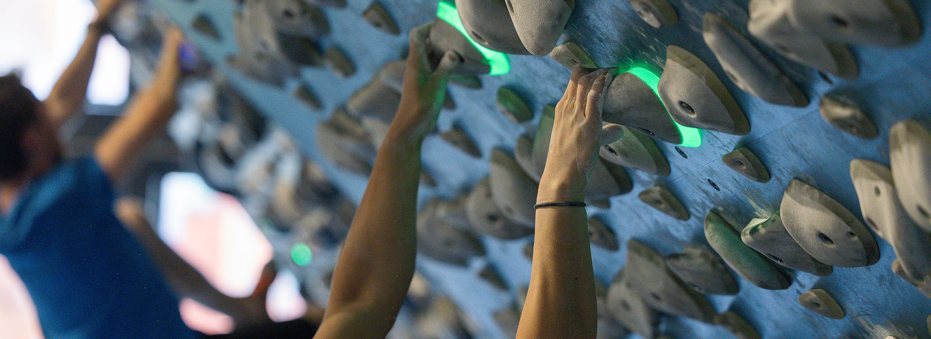 Close-up of people climbing an indoor rock wall, focusing on hands gripping illuminated green climbing holds and poplar training boards, with blurred climbers and holds in the background.