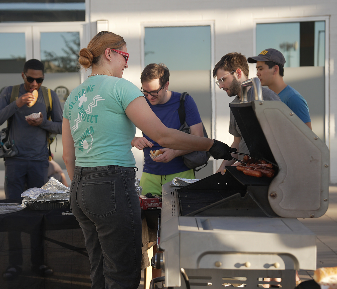 A woman grills sausages on an outdoor barbecue while four men stand nearby, some eating and chatting. The group appears to be enjoying a casual Climb for Community gathering in front of a modern building.