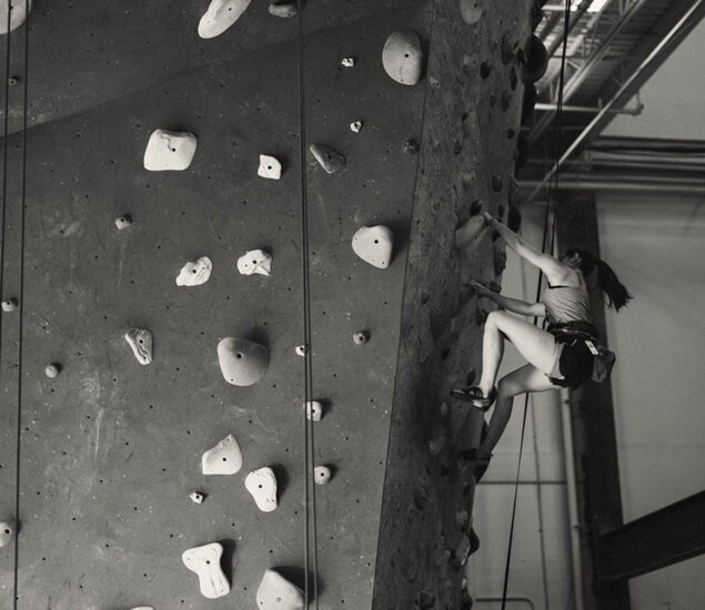 A person wearing a harness climbs an indoor rock climbing wall, gripping handholds and footholds, with ropes for safety. The black-and-white scene captures the determination often seen in Seattle youth teams as they train and scale new heights.