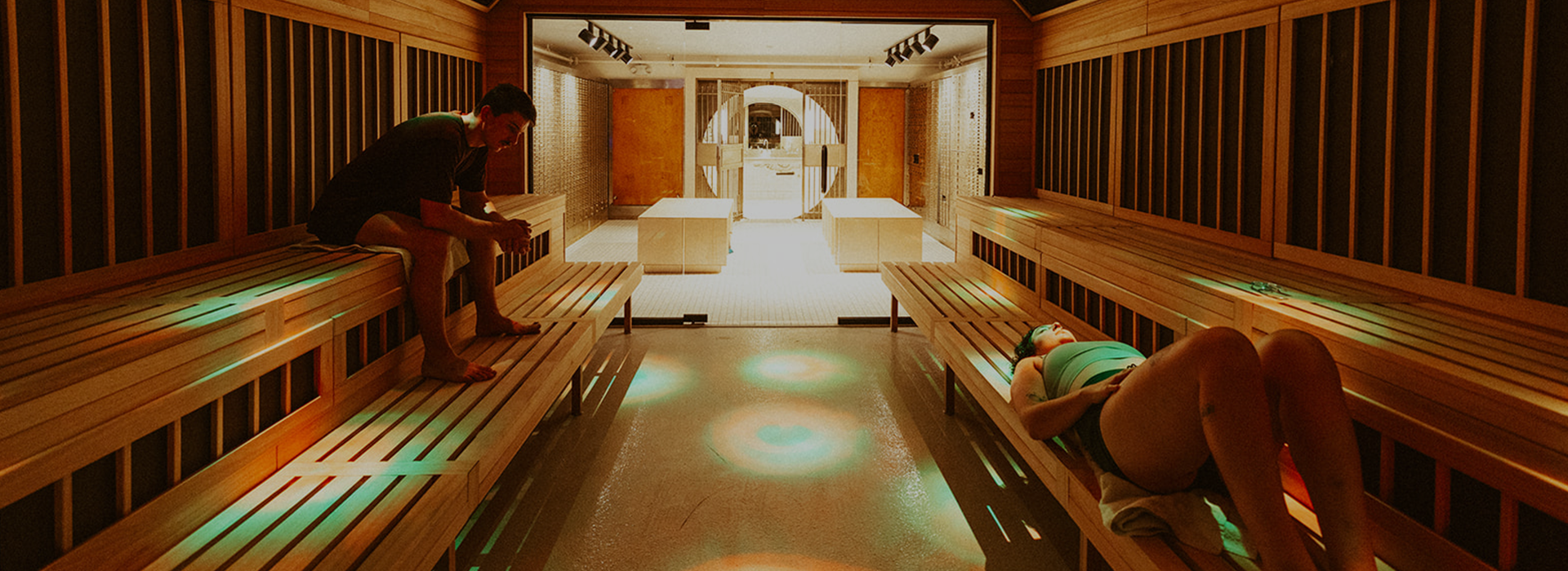 Two people relax on wooden benches inside a spacious, modern sauna with soft, colorful lighting and a bright central entryway—one of the many membership benefits at Seattle Bouldering Project.