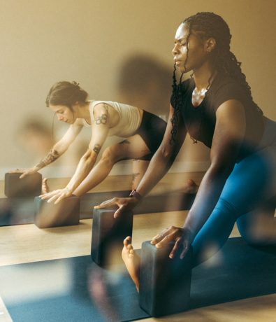 Two women practicing yoga indoors, each in a low lunge pose using yoga blocks on mats. They look focused and calm, enjoying natural light—an example of the membership benefits at Seattle Bouldering Project.