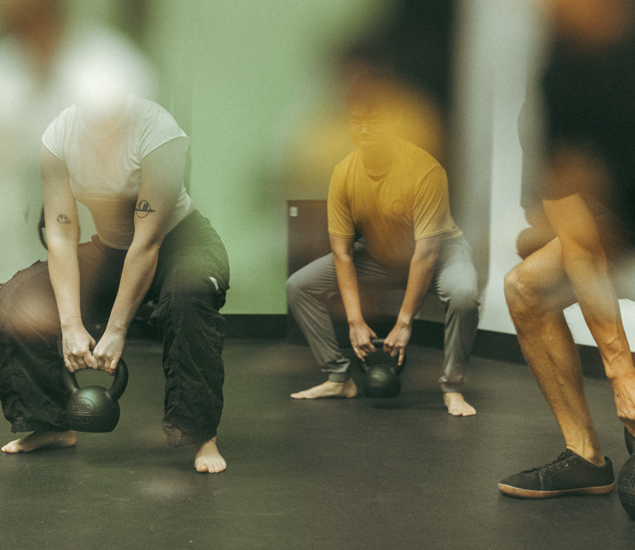 A group of people exercise indoors at Seattle Bouldering Project, performing squats with kettlebells. Barefoot or in minimal footwear, they enjoy membership benefits as the photo features a soft, blurred effect in some areas.