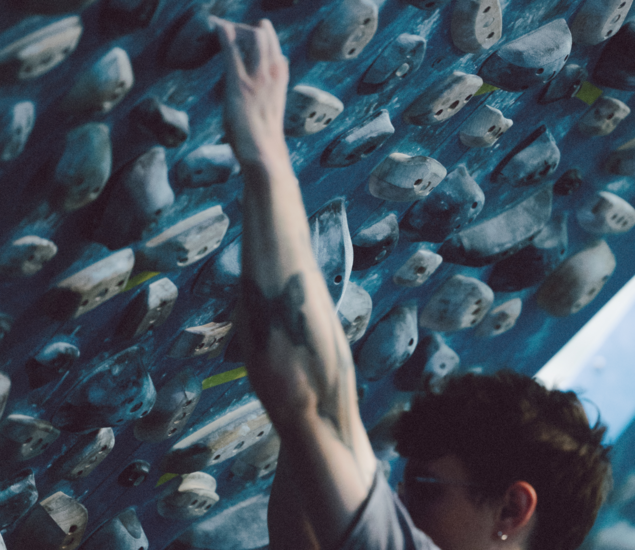 A person with tattoos on their arm reaches for a hold on an indoor climbing wall covered with poplar training boards and various handholds. The setting appears to be a bouldering gym.