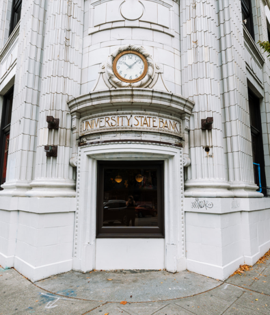 Ornate white building corner with a clock showing 12:05 above an arched “University State Bank” sign, and a window below reflecting the street and photographer—like glimpsing Membership Benefits at Se