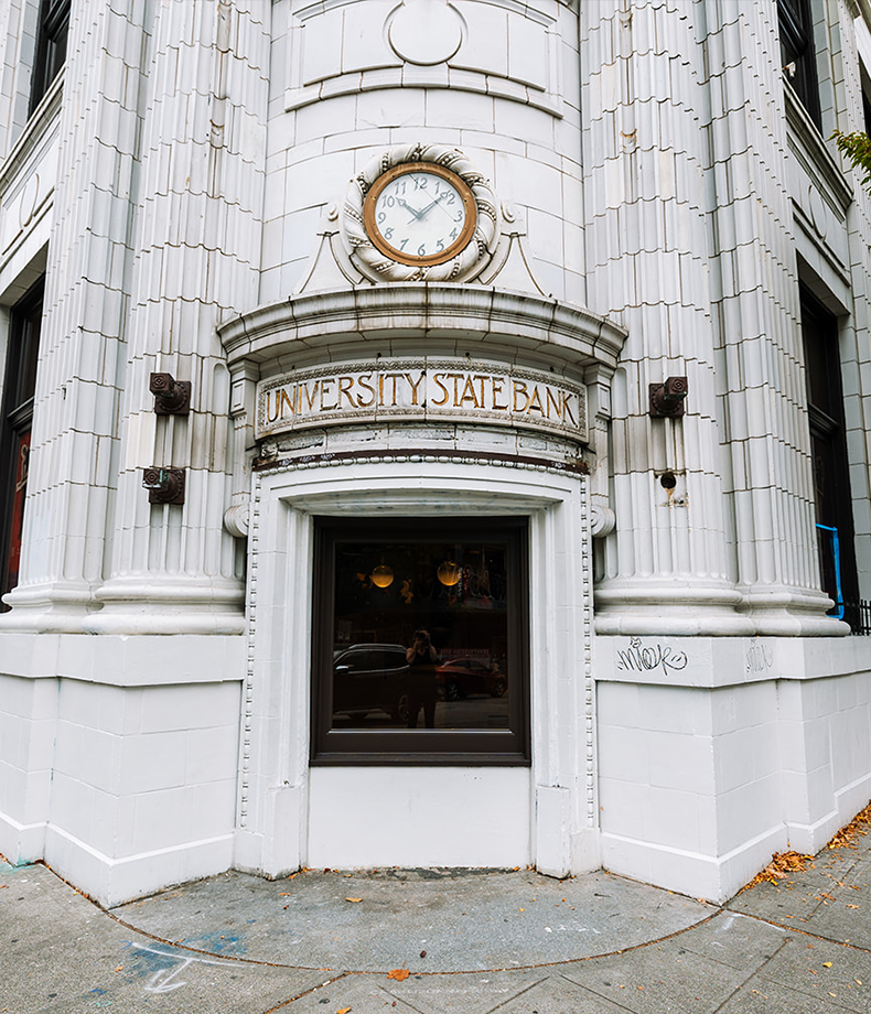 Ornate white building corner with a clock showing 12:05 above an arched “University State Bank” sign, and a window below reflecting the street and photographer—like glimpsing Membership Benefits at Se