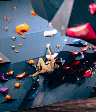 A person climbs an indoor bouldering wall with colorful handholds and footholds, wearing casual clothes and climbing shoes. The wall has blue panels and angled surfaces—just one perk among the Membership Benefits at Seattle Bouldering Project.