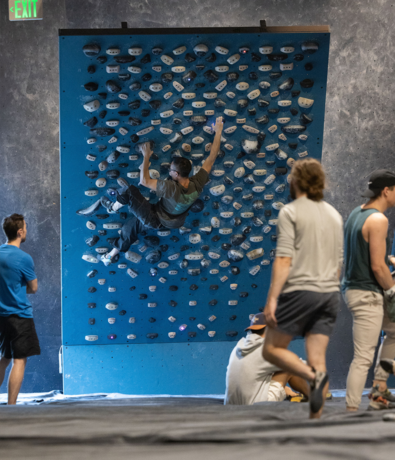 A person climbs a blue indoor bouldering wall with many handholds, while several people watch or wait nearby on padded flooring—enjoying the Membership Benefits at Seattle Bouldering Project in this vibrant climbing gym.