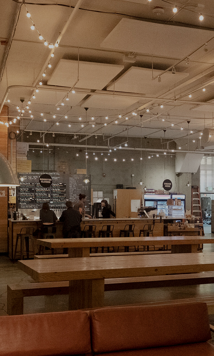 A cozy, modern café with string lights on the ceiling, a wooden bar, chalkboard menus, and people at the counter. Long wooden tables fill the foreground—perfect for chatting about Membership Benefits at Seattle Bouldering Project.