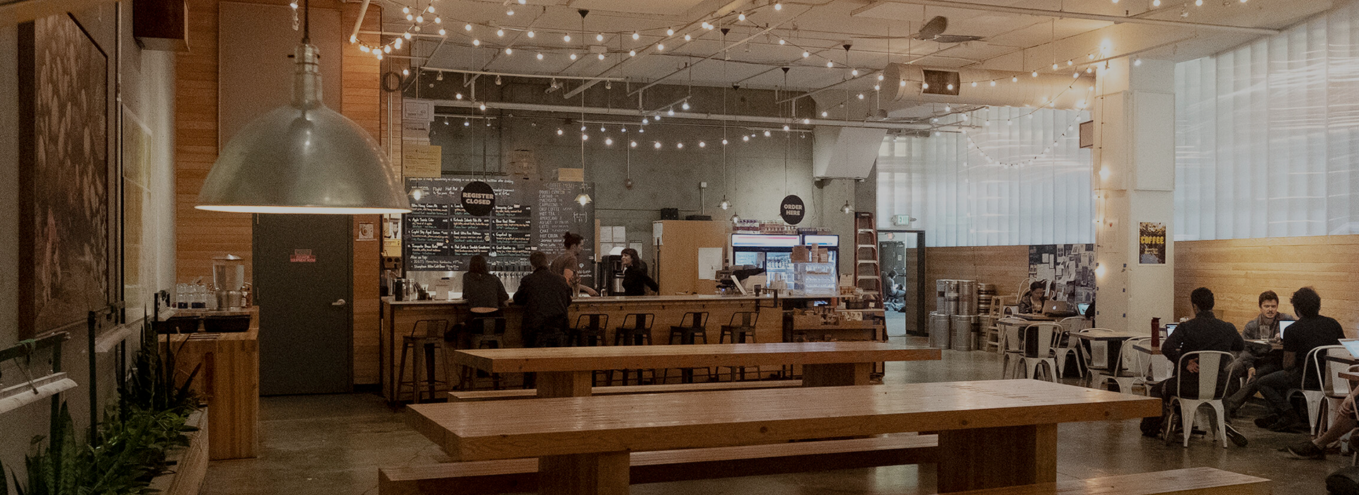 A modern coffee shop with long wooden tables, hanging string lights, a bar counter with stools, and people chatting in a cozy space—perfect for discussing Membership Benefits at Seattle Bouldering Project in an inviting, industrial-style setting.