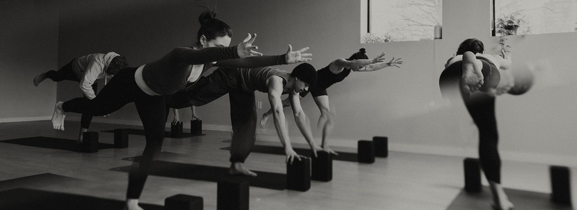 A group of people practicing yoga in a studio at Seattle Bouldering Project, all balancing on one leg with arms extended forward, using yoga blocks for support. Large windows and a calm atmosphere highlight the membership benefits offered here.