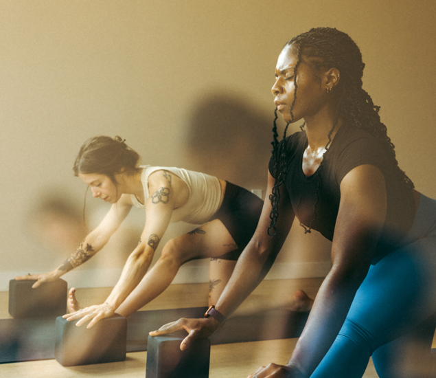 Two women in athletic wear practice yoga indoors, using yoga blocks for support on yoga mats. They appear focused and calm in a softly lit room, enjoying Membership Benefits at Seattle Bouldering Project.