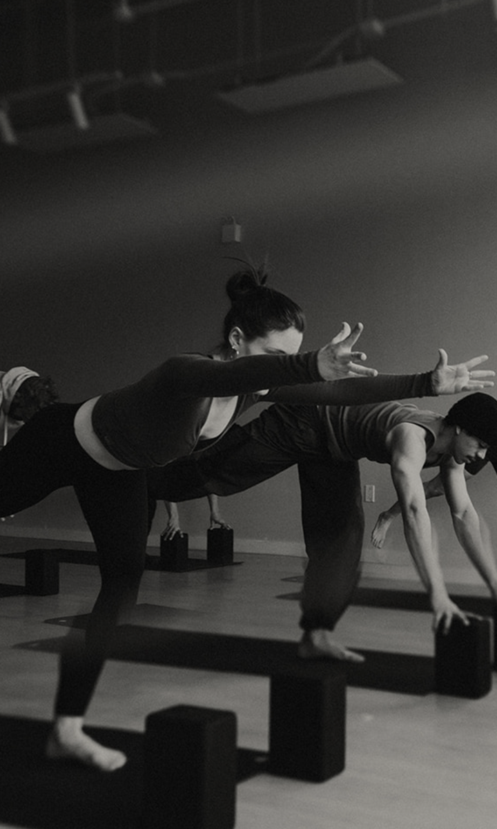 A black-and-white photo of people in a yoga class performing a balancing pose on one leg, with their arms extended forward and yoga blocks on the floor—highlighting Membership Benefits at Seattle Bouldering Project.