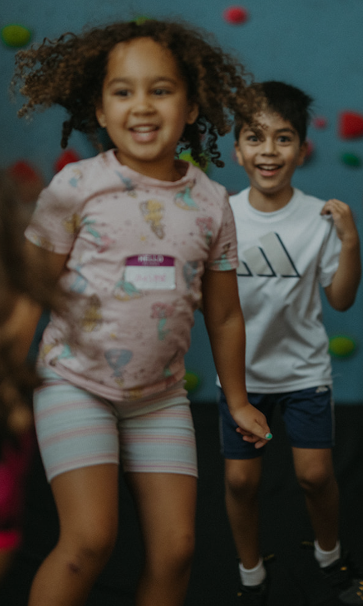 Two children are smiling and playing indoors in front of a blue wall with colorful climbing holds. Their joyful energy highlights the fun and Membership Benefits at Seattle Bouldering Project, where young climbers thrive together.