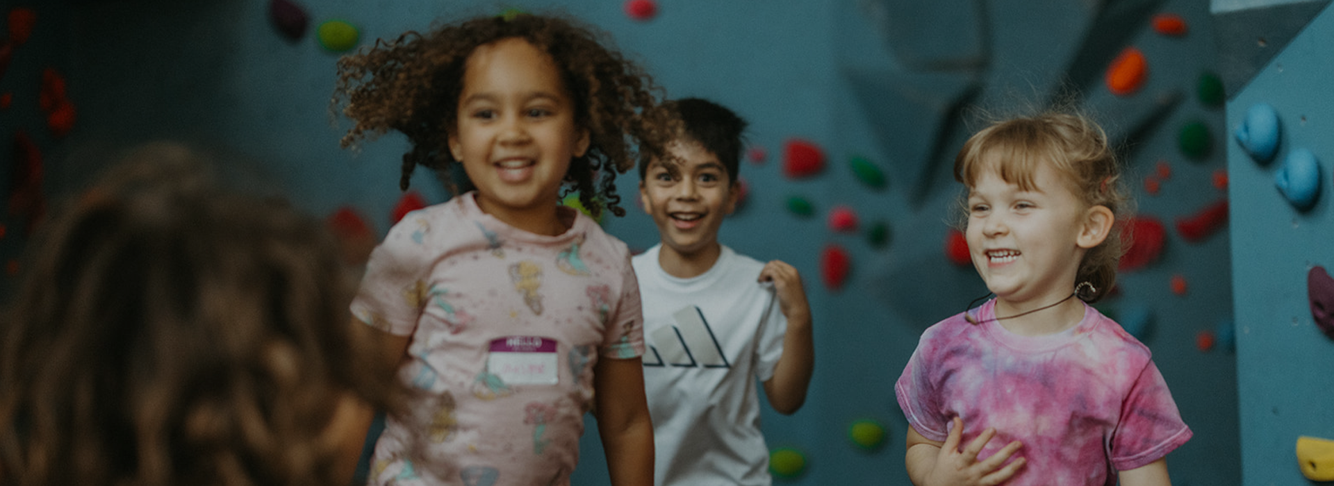 Three young children smile and laugh together indoors, standing in front of a colorful climbing wall at Seattle Bouldering Project, where membership benefits offer families access to creative play and climbing fun.