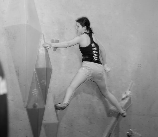 A woman in a tank top and shorts climbs a bouldering wall in Seattle, skillfully balancing on large, angular holds. The black and white image captures the determination often seen in local youth teams.