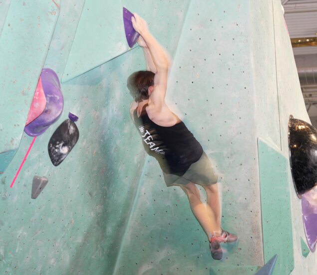A person wearing a black tank top and shorts climbs an indoor bouldering wall in Seattle, gripping large purple holds with both hands and lifting their body off the ground—perfect training for local youth teams.