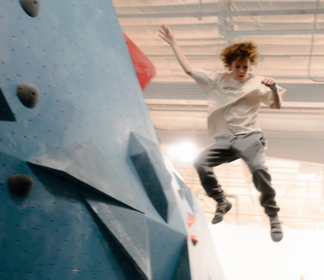 A young member of Seattle Youth Teams with curly hair and casual clothes is jumping off an indoor climbing wall, mid-air, arms outstretched and intense expression, in a brightly lit gym.