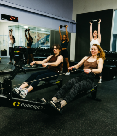 Four women exercising in a Boston climbing gym; two use rowing machines in the foreground while two others lift dumbbells with raised arms behind them. A wall clock and mirrors are visible. Perfect spot to start your summer membership!.