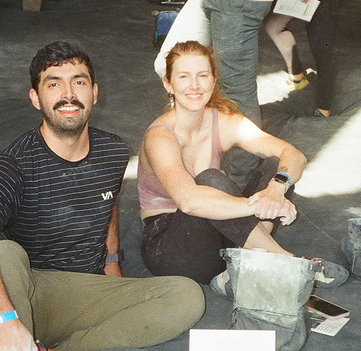 Two people are sitting on the floor of an indoor space, smiling at the camera during a Seattle climbing competition. Climbing gear and chalk bags are nearby, while others in the background enjoy the sunlight streaming across the floor.