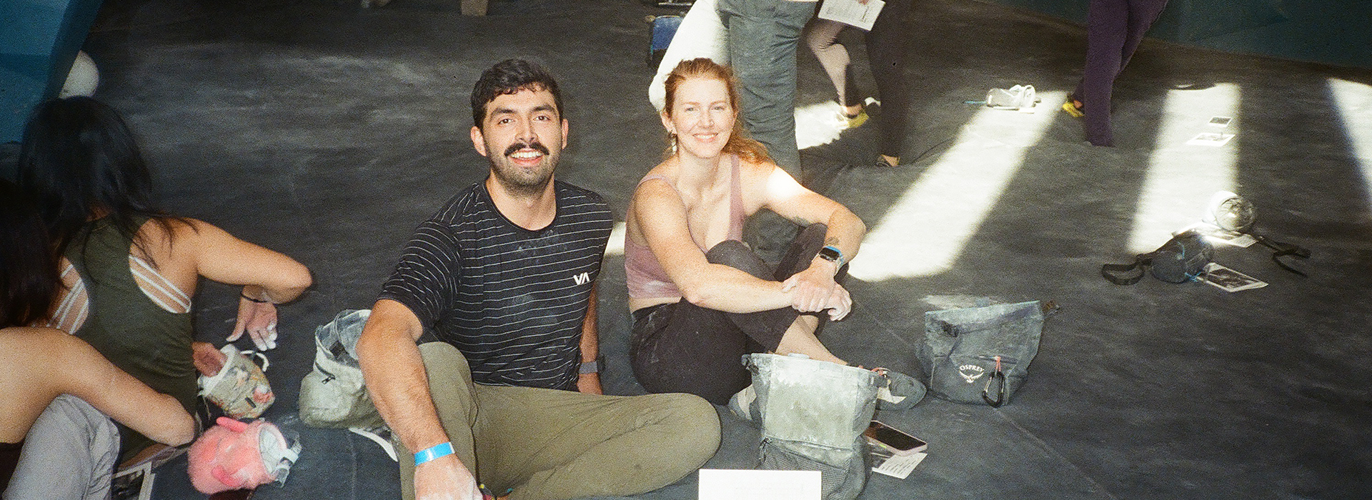 Two people are sitting on the floor of an indoor space, smiling at the camera during a Seattle climbing competition. Climbing gear and chalk bags are nearby, while others in the background enjoy the sunlight streaming across the floor.