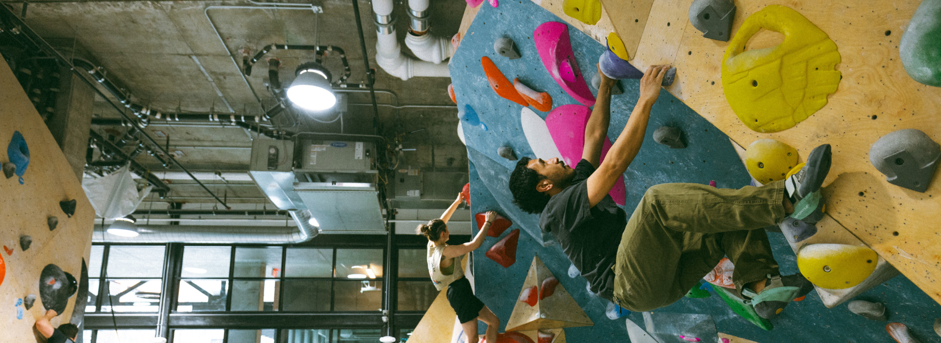 Two people climb an indoor bouldering wall with colorful holds at a DC climbing gym. The space features an industrial design, with visible pipes, ducts, and large windows in the background.