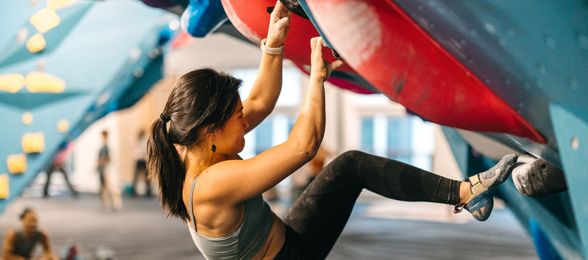 A woman in athletic wear climbs an indoor bouldering wall, gripping large red holds and using her legs for support—a great showcase of rock climbing muscles in action. The gym environment is visible in the background.