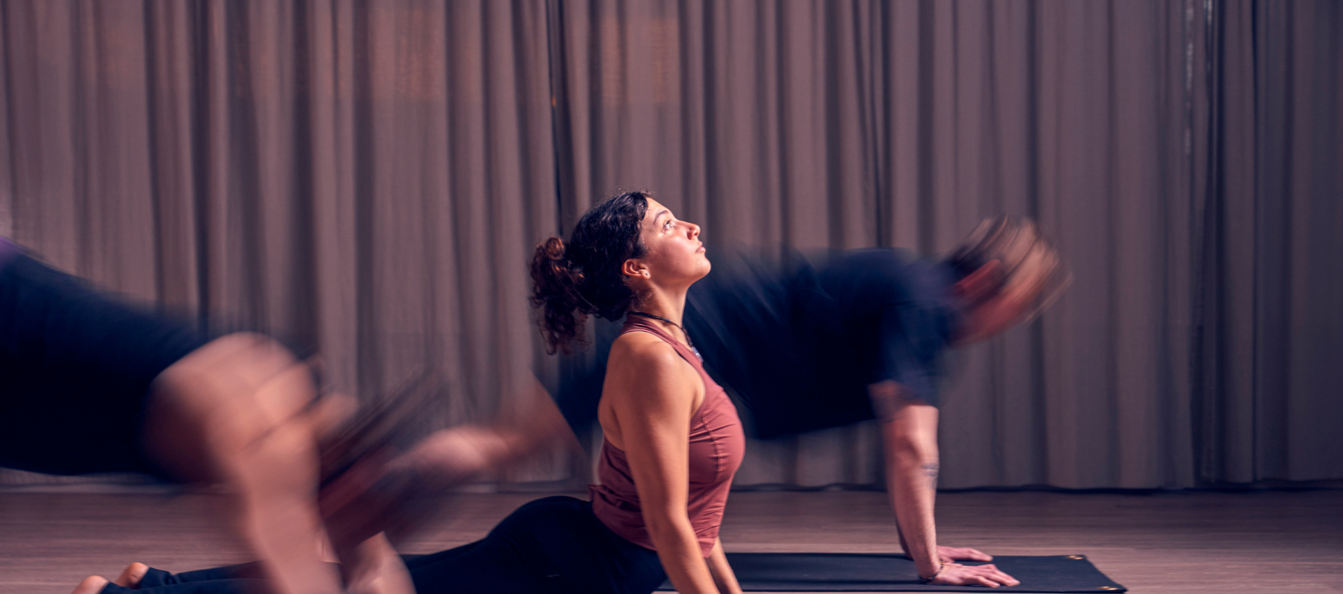 A woman in a yoga studio does an upward-facing dog pose on a mat, looking upward—an ideal move to improve mobility and strength. Two blurred figures nearby suggest movement as they transition between poses. A neutral curtain hangs in the background.