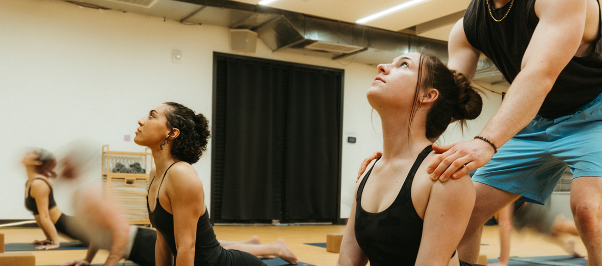 Two women in a yoga class perform a seated twist, one of the best yoga poses for climbers, as an instructor in blue shorts gently guides them. The studio features wooden floors and a high ceiling with exposed ducts.