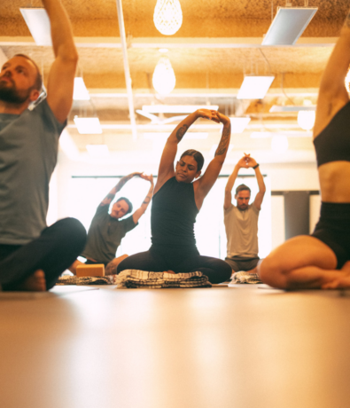 A group of people sit cross-legged on mats in a bright room at a Boston climbing gym, stretching their arms overhead during a yoga or meditation class. The focus is on one person in the center, with others mirroring the pose—perfect for your summer membership.