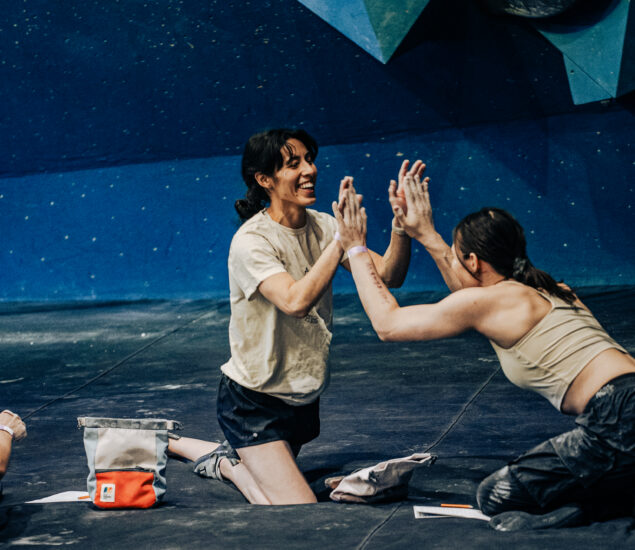 Two women wearing athletic clothing and covered in chalk sit on the gym floor, smiling and clapping hands in celebration after a seattle climbing competition, with climbing walls and gear visible in the background.