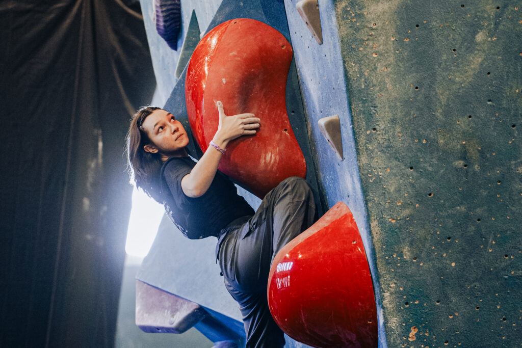 A person with long hair climbs an indoor bouldering wall, gripping large red holds and looking upward with determination. The blue wall features various textured and triangular holds, making bouldering a full body workout.