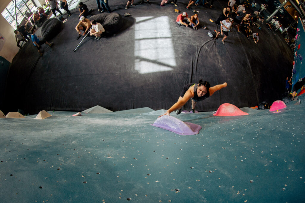 A person is climbing an indoor bouldering wall, reaching for a hold, while others watch or rest on mats below in a busy gym filled with bright, natural light—a lively scene from any beginner’s guide to bouldering.