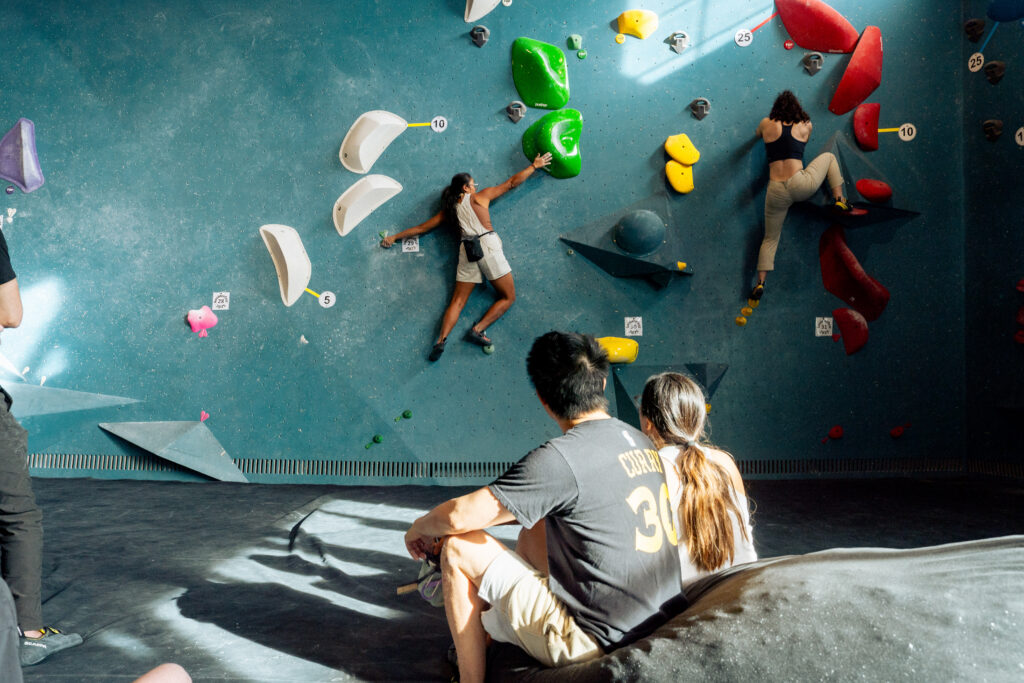 Two people are indoor rock climbing on a blue wall with colorful holds, while a man and woman sit on the floor watching them. Sunlight streams in, casting shadows—an inspiring scene for anyone exploring a beginner’s guide to bouldering.