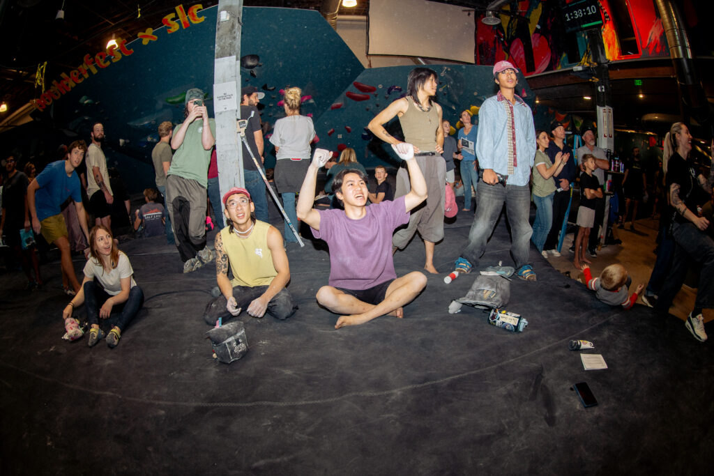 A group at an indoor rock climbing gym, some sitting and some standing, watch an event. One person in a purple shirt sits cross-legged, raising their arms in excitement—a lively moment perfect for a beginner’s guide to bouldering.