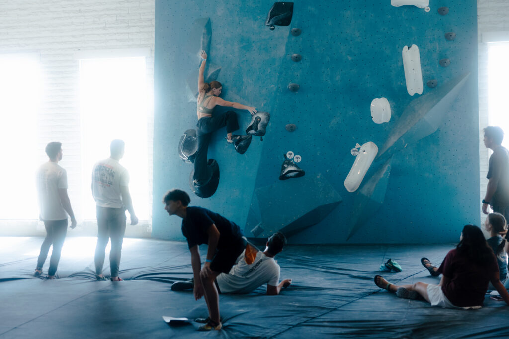 A group of people watch as a woman climbs a blue indoor bouldering wall, reaching for a hold. Some spectators stand, while others sit or kneel on padded mats below. Bouldering offers one of the best full body workouts as bright light streams in from tall windows.