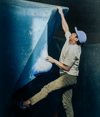 A person wearing a cap and climbing shoes reaches up with one hand while bouldering on a large blue indoor wall at a Boston climbing gym, enjoying the challenge and perks of their summer membership.
