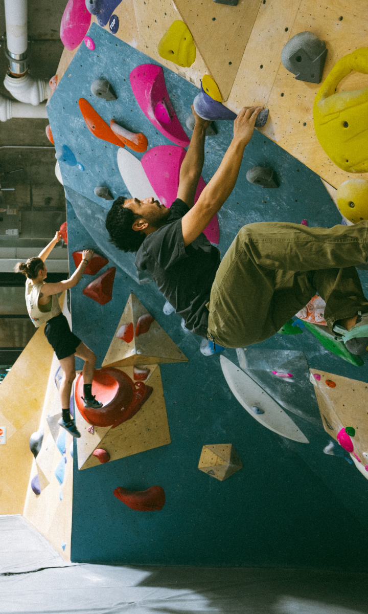 Two people are indoor rock climbing at a DC climbing gym on a colorful bouldering wall; one scales vertically in the background, while the other tackles a steep overhang in the foreground, gripping vibrant handholds.