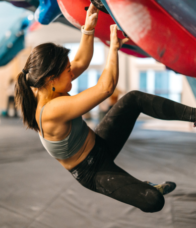 A woman in athletic clothing climbs an indoor bouldering wall, gripping red holds as she engages key rock climbing muscles for support. Focused and suspended above padded flooring, she demonstrates a challenging rock climbing workout.