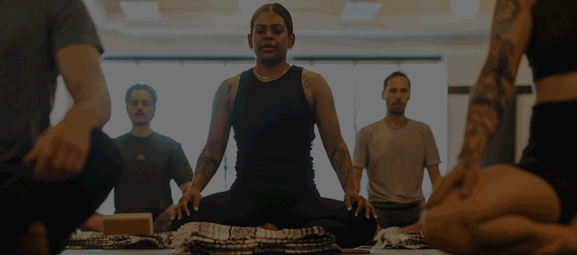 A group of people sitting cross-legged on yoga mats in a bright room, eyes closed and hands resting on their knees, enjoying a peaceful meditation or yoga session as part of their summer membership.