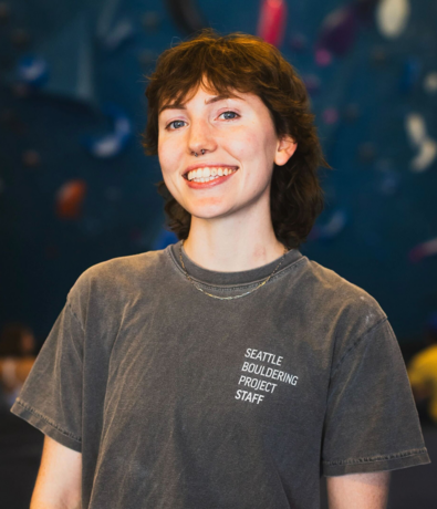 A person with short brown hair and a nose ring smiles at the camera, wearing a gray Seattle Bouldering Project Staff t-shirt. The blurred background hints at climbing holds, capturing the spirit of summer membership.