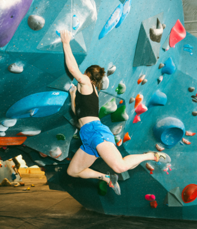 A person in blue shorts and a black tank top is climbing a colorful indoor bouldering wall, gripping holds with one hand while their body extends outward. Bouldering is known as one of the best workouts for a full body workout.