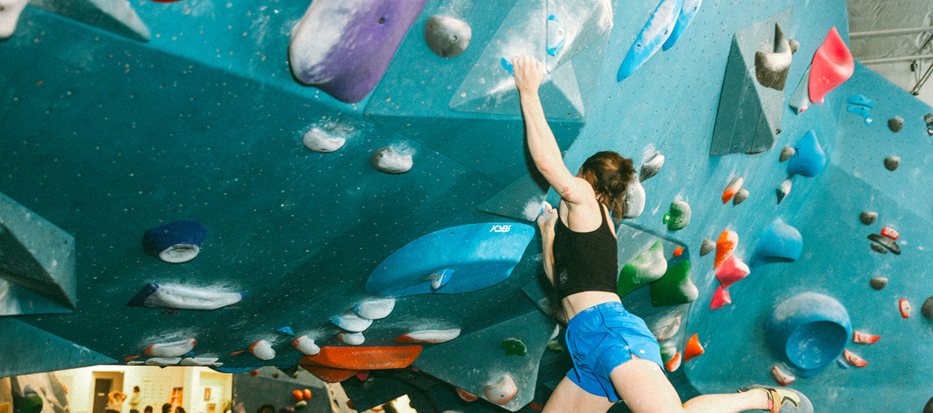 A person wearing a black tank top and blue shorts climbs an indoor bouldering wall covered in colorful holds, reaching with one arm toward the next grip—showcasing one of the best full body workouts.